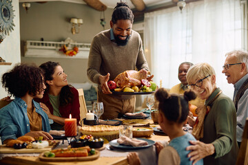 Happy black man serving Thanksgiving turkey to his extended family at dining table.