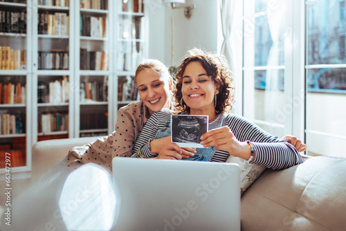 Happy young lesbian couple showing the x-ray of their future newborn on the laptop video call at home photo