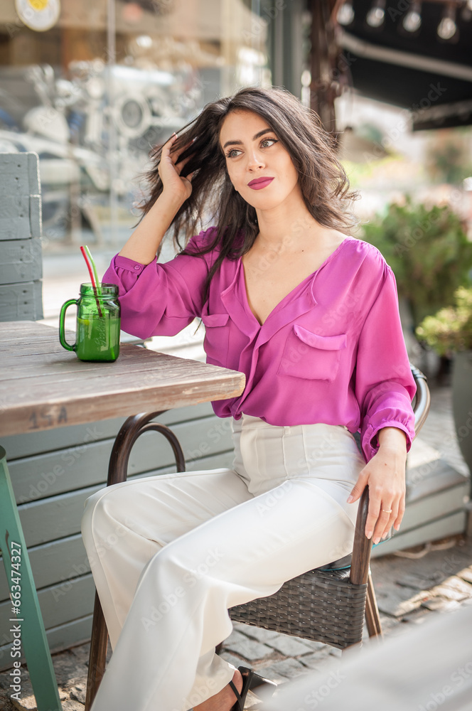 Happy Brunette Girl with pink blouse Sitting at the bar , Drinking a glass of lemonade While Smiling at the camera. Young sexy pretty woman on the chair drinking juice wearing a white pants