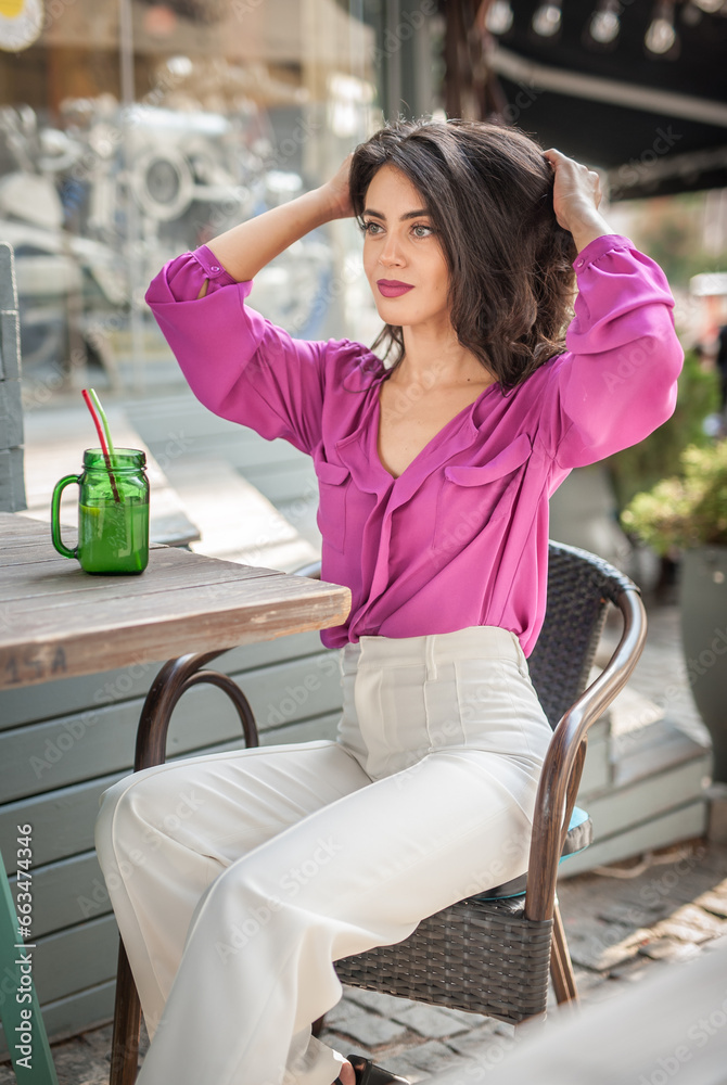 Happy Brunette Girl with pink blouse Sitting at the bar , Drinking a glass of lemonade While Smiling at the camera. Young sexy pretty woman on the chair drinking juice wearing a white pants