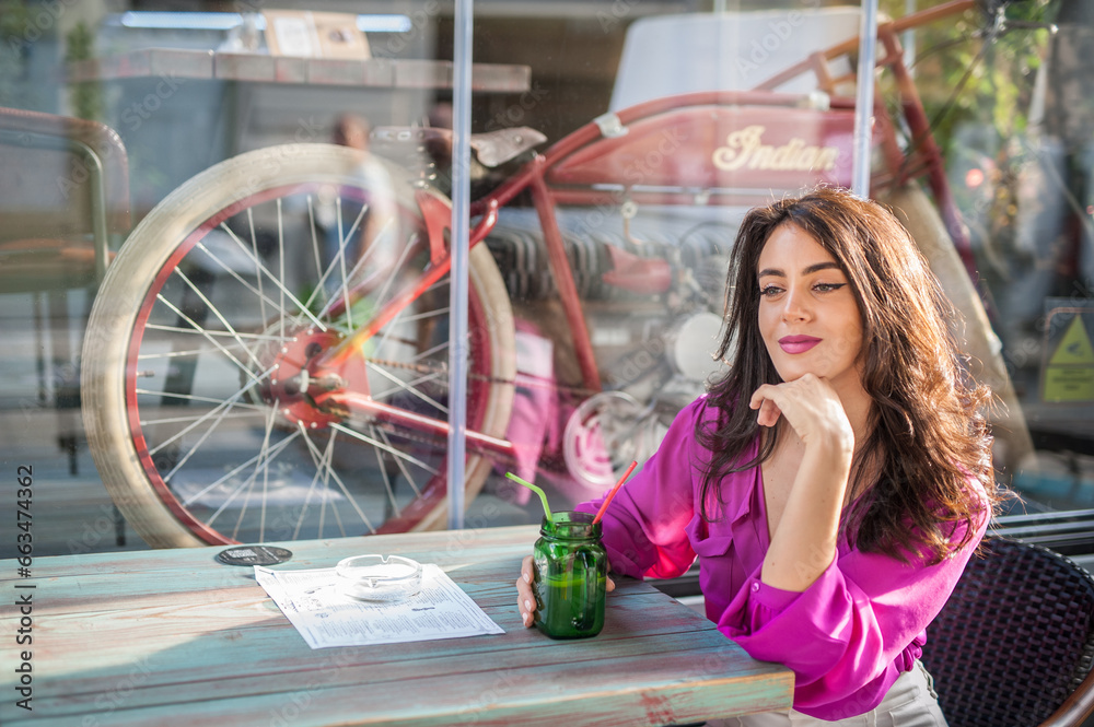 Happy Brunette Girl with pink blouse Sitting at the bar , Drinking a glass of lemonade While Smiling at the camera. Young sexy pretty woman on the chair drinking juice wearing a white pants