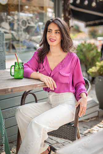 Happy Brunette Girl with pink blouse Sitting at the bar , Drinking a glass of lemonade While Smiling at the camera. Young sexy pretty woman on the chair  drinking  juice wearing a white pants