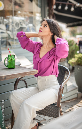 Obraz na płótnie Happy Brunette Girl with pink blouse Sitting at the bar , Drinking a glass of lemonade While Smiling at the camera. Young sexy pretty woman on the chair drinking juice wearing a white pants w salonie