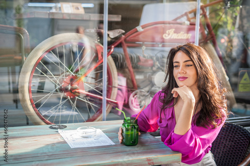 Obraz na płótnie Happy Brunette Girl with pink blouse Sitting at the bar , Drinking a glass of lemonade While Smiling at the camera. Young sexy pretty woman on the chair drinking juice wearing a white pants w salonie
