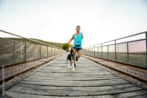 Frontal photo of an adult runner with an amputated arm running on an outdoor bridge with a dog attached to a harness. Canicross concept. Activities with a border collie. Running with pets.