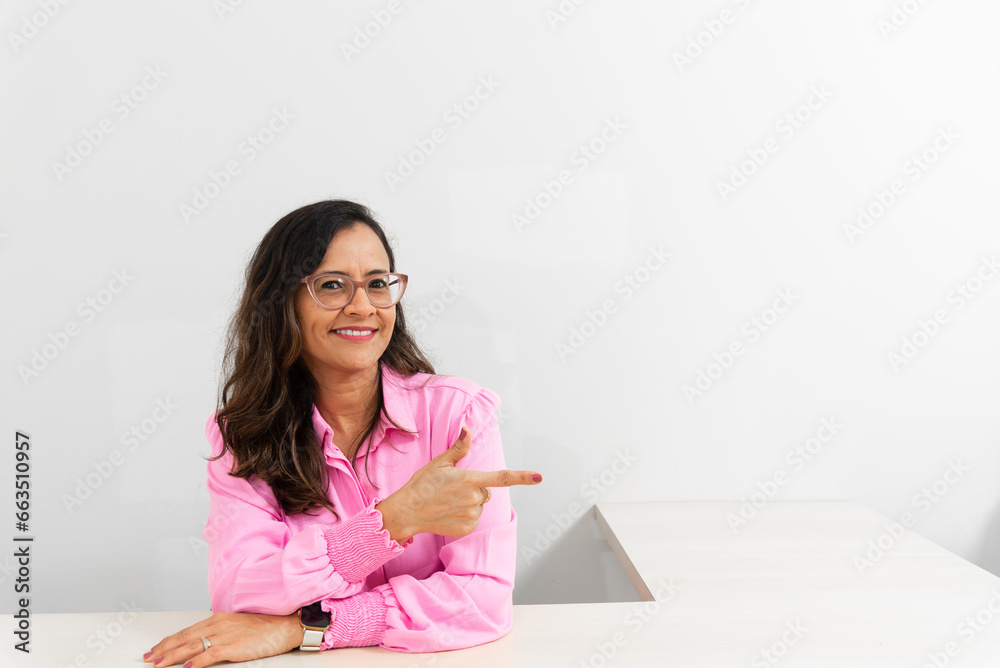 Portrait of a psychopedagogue, wearing pink clothes, in her office, sitting, making a sign with her hand. Helps with school performance. Portrait of a psychopedagogue, wearing pink clothes, in her office, sitting, making a sign with her hand. Helps with school performance.