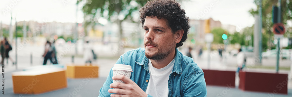 Close-up portrait of young bearded man in denim shirt sitting in wireless headphones and drinking coffee on busy street modern city background. Profile of man drinking coffee Close-up portrait of young bearded man in denim shirt sitting in wireless headphones and drinking coffee on busy street modern city background. Profile of man drinking coffee