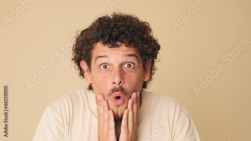 Close up, shocked guy with curly hair dressed in beige t-shirt looking in surprise at the camera with big eyes, raises his hands to his face, wow, isolated on beige background in the studio