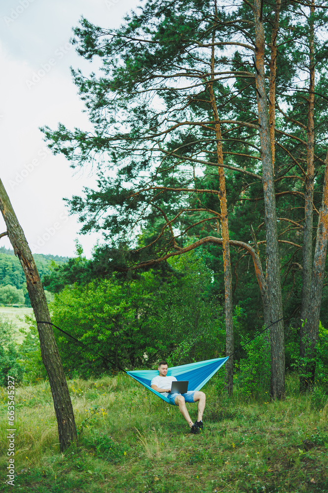A young man with a laptop sits in a hammock in nature and works remotely. Work in nature during vacation. A young man with a laptop sits in a hammock in nature and works remotely. Work in nature during vacation.