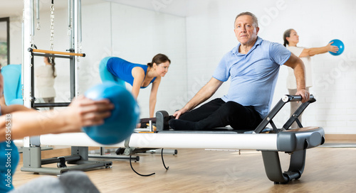 Diligent elderly man practicing pilates on reformer tower equipment in sports hall during pilates classes. Persons doing pilates with trainer