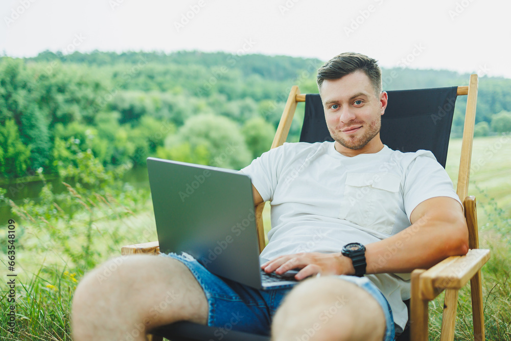 Obraz premium View of handsome guy lying on chair in nature and working with laptop, resting alone, thoughtfully looking up. A man on a trip, in the countryside. the concept of people's lifestyle