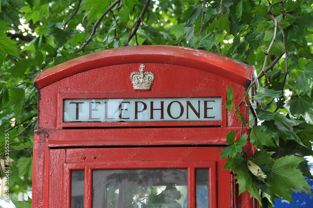The top of a classical British red telephone booth surrounded by green maple leaves. The top of a classical British red telephone booth surrounded by green maple leaves.
