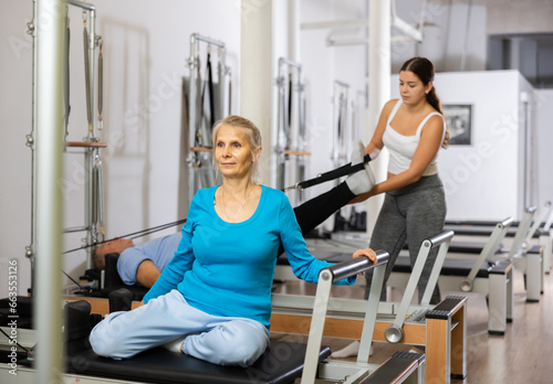 Positive elderly woman in sportswear getting ready for performing torso rotation on Pilates performer bed machine in rehabilitation center