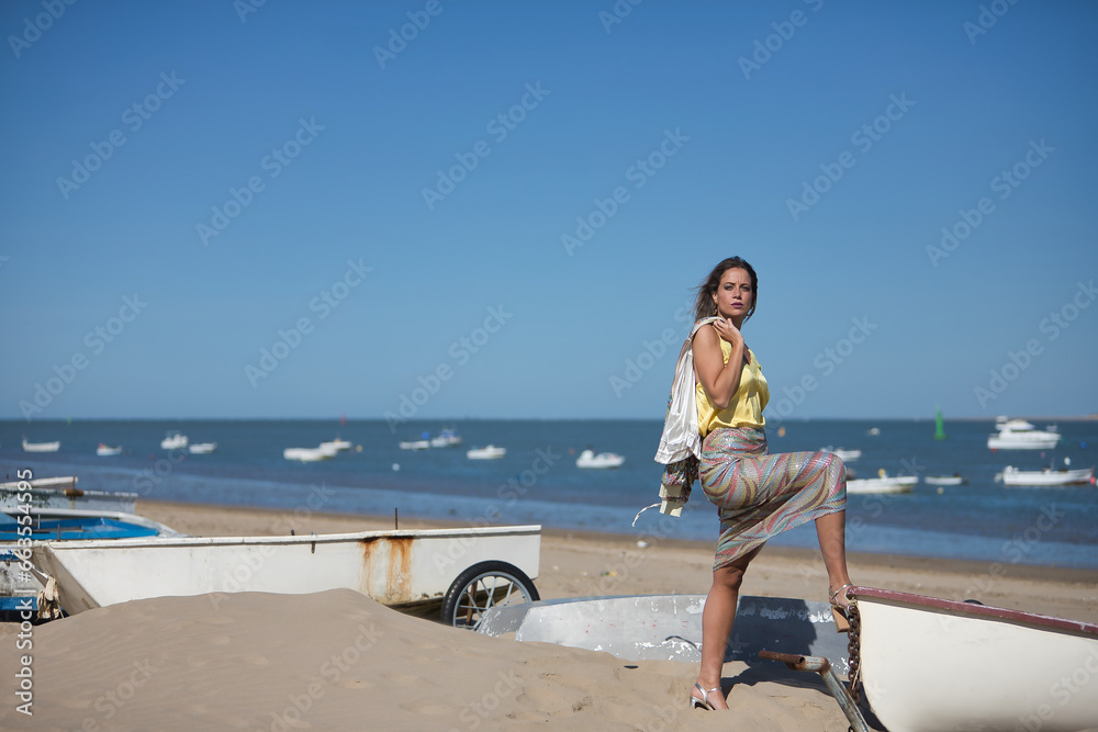 Young and beautiful woman in a sequined dress and high heels, with her foot on a boat on the beach, posing looking at the camera. Concept beauty, fashion, trend, empowerment.