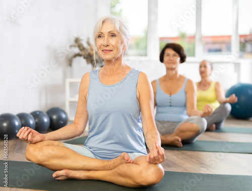 Positive old woman practicing lotus pose of yoga on black rug during fitness classes