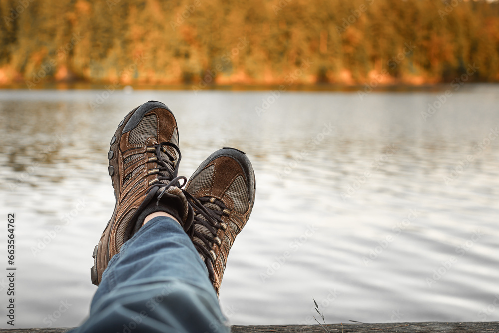 Man with hiking shoes relaxing by an autumn mountain lake. Adventure and people finding peace alone in nature  - obrazy, fototapety, plakaty 