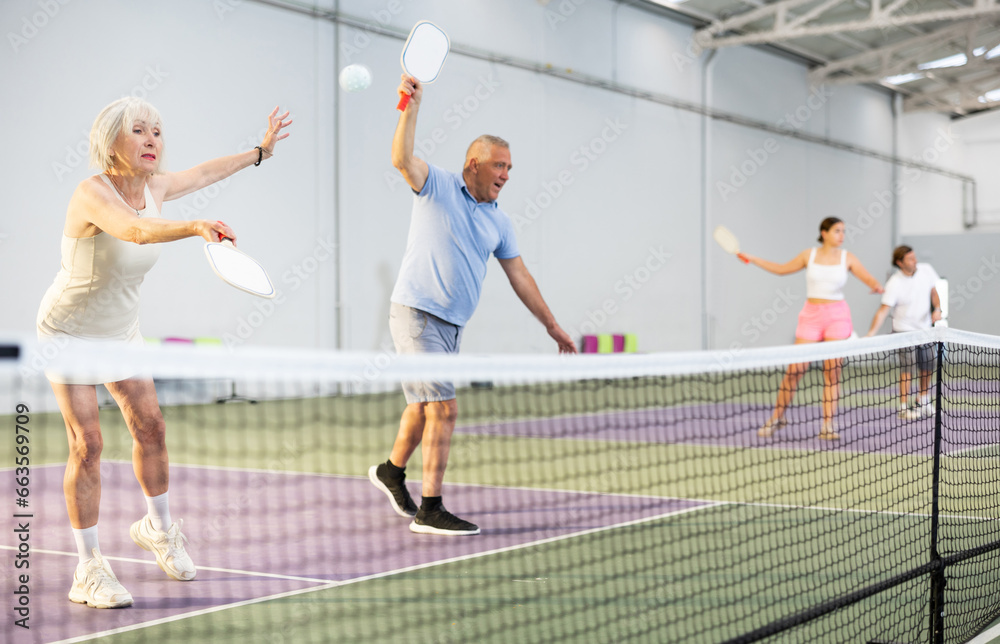Focused positive elderly woman playing friendly pickleball match in team with male partner on indoors court. Concept of concentration in competition