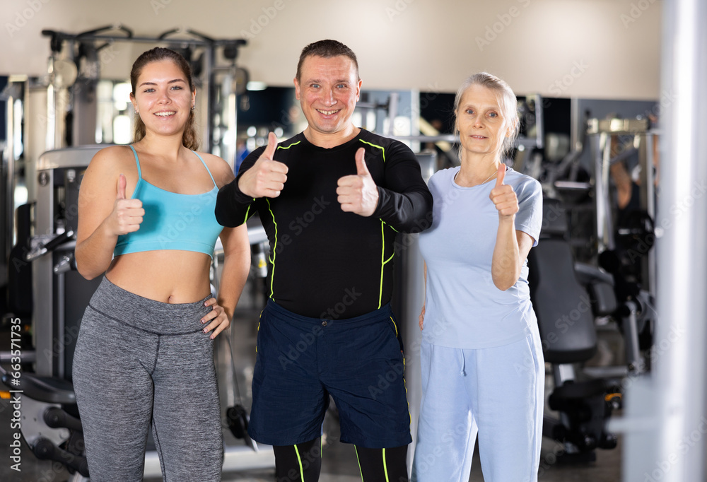 Portrait of cheerful sporty people of different ages standing in modern gym during workout, showing thumbs up. Concept of popularity of active healthy lifestyle