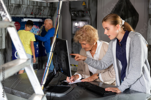 Senior woman and her granddaughter using computer to solve riddle in escape room. Young boy and grandfather standing in background and looking at map on wall.