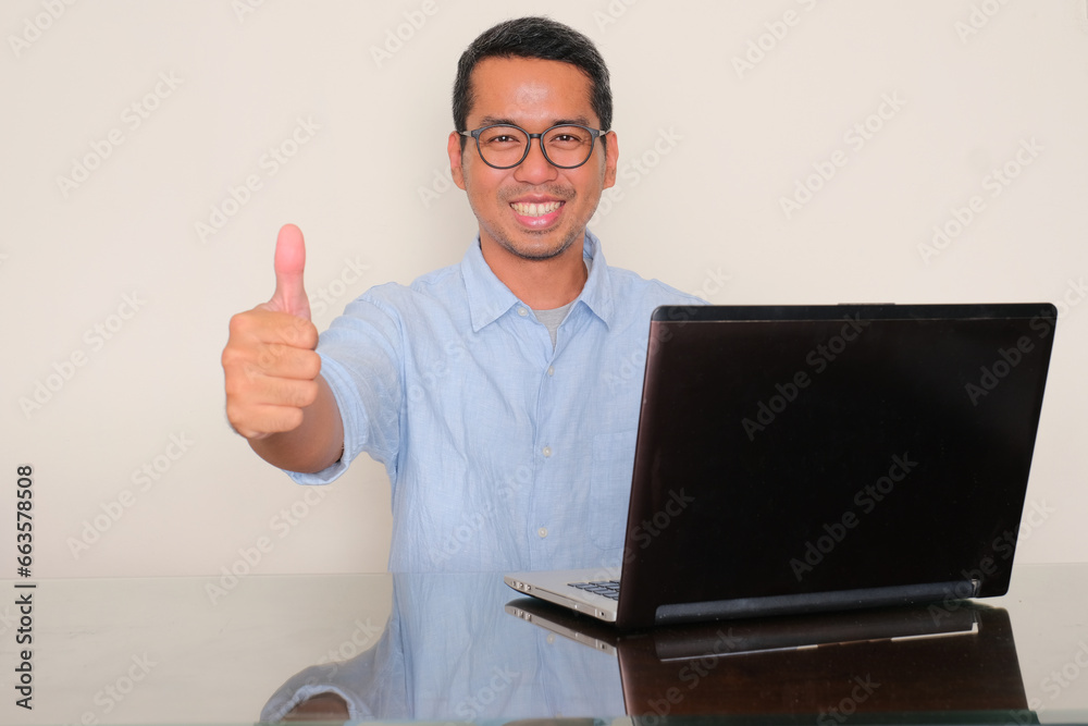 Adult Asian man sitting in front of a laptop smiling and give thumb up Adult Asian man sitting in front of a laptop smiling and give thumb up