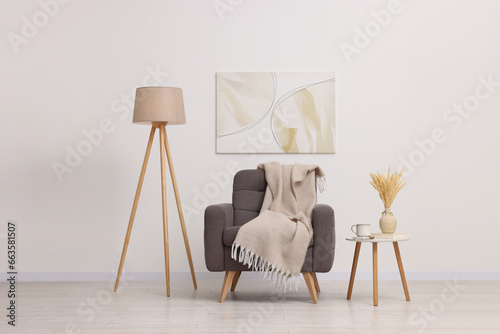 Comfortable armchair, blanket, lamp and side table near white wall indoors photo