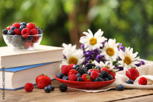 Different fresh ripe berries, beautiful flowers and books on wooden table outdoors