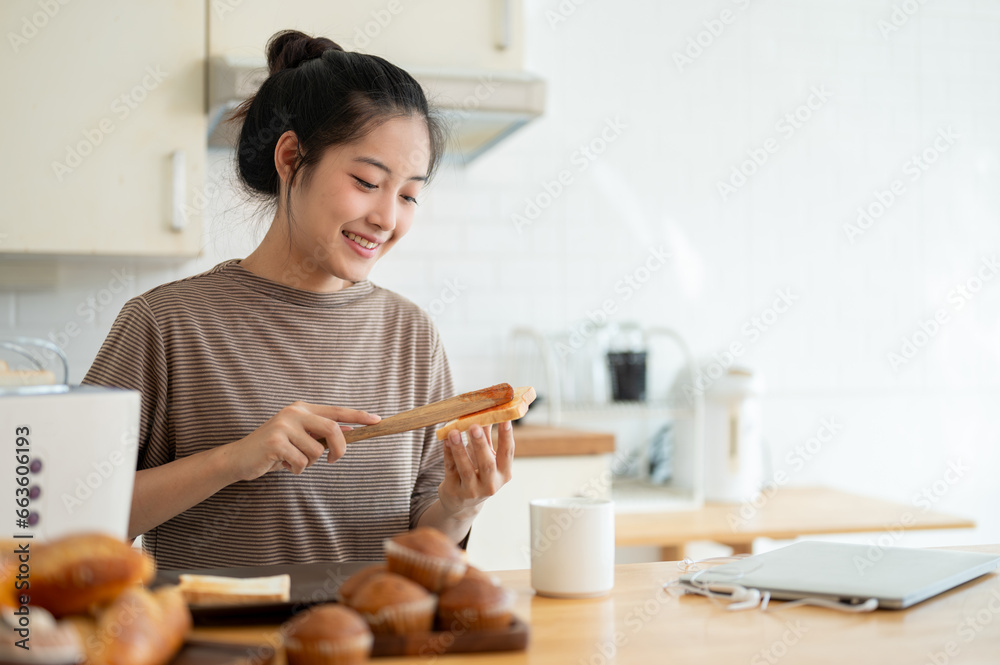 A pretty Asian woman is spreading strawberry jam onto a toast, preparing her breakfast in the kitchen