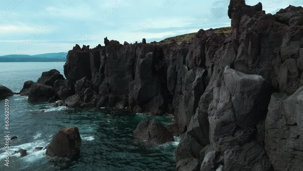 Top view of black rocks of sea coast on cloudy day. Clip. Sheer black rocks on shore of northern ocean on cloudy summer day. Cinematic landscape of black cliff on ocean coast