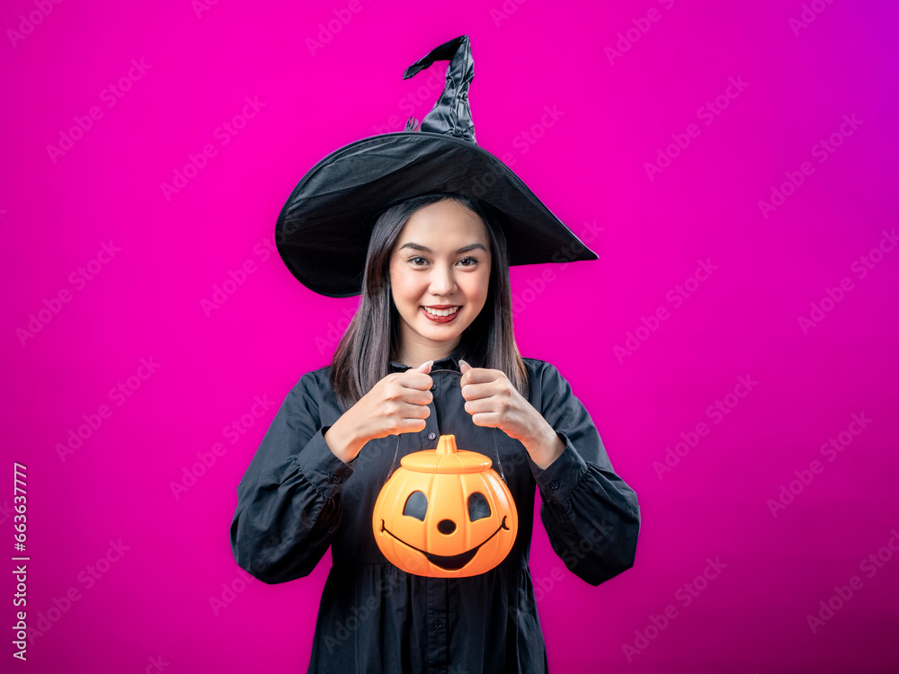 Portrait of an Asian Indonesian woman wearing a Halloween-themed costume with a witch hat, smiling while holding a pumpkin. Isolated against a magenta background. Portrait of an Asian Indonesian woman wearing a Halloween-themed costume with a witch hat, smiling while holding a pumpkin. Isolated against a magenta background.