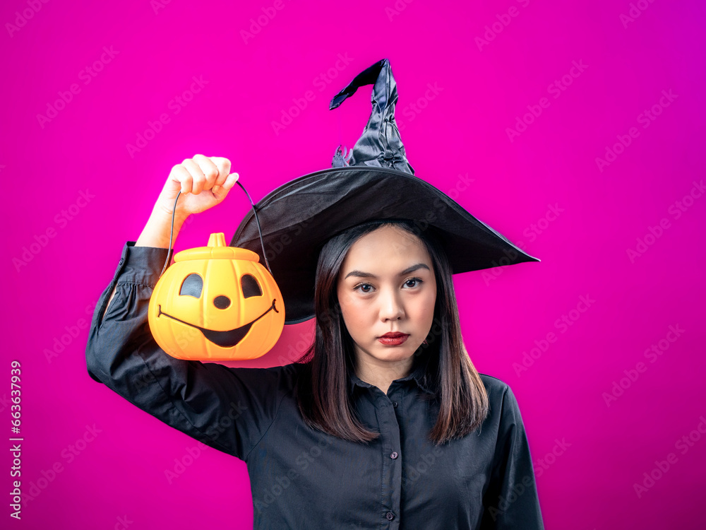 Portrait of an Asian Indonesian woman wearing a Halloween-themed costume with a witch hat, smiling while holding a pumpkin. Isolated against a magenta background. Portrait of an Asian Indonesian woman wearing a Halloween-themed costume with a witch hat, smiling while holding a pumpkin. Isolated against a magenta background.