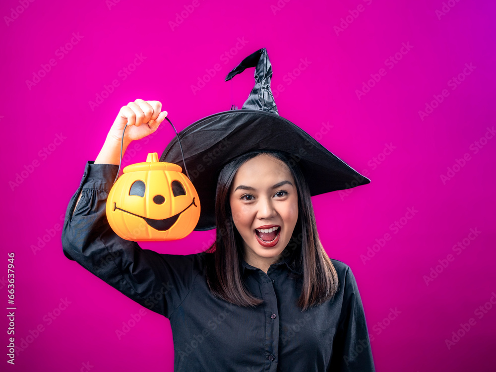 Portrait of an Asian Indonesian woman wearing a Halloween-themed costume with a witch hat, smiling while holding a pumpkin. Isolated against a magenta background. Portrait of an Asian Indonesian woman wearing a Halloween-themed costume with a witch hat, smiling while holding a pumpkin. Isolated against a magenta background.