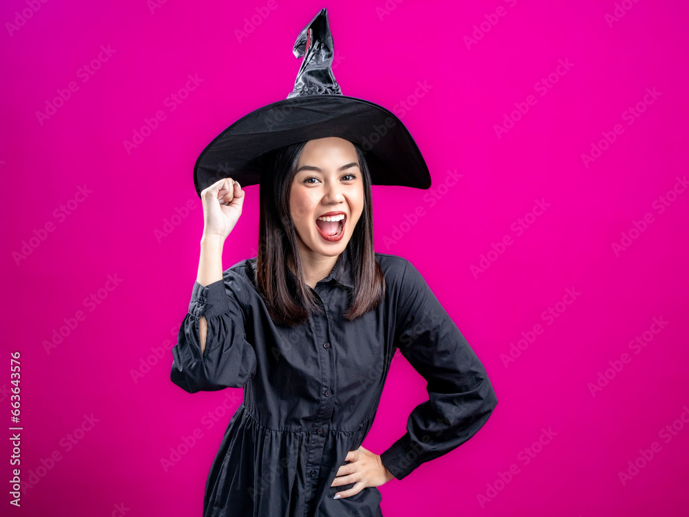 Portrait of an Asian Indonesian woman wearing a Halloween-themed costume with a witch hat, celebrating with clenched fists. Isolated against a magenta background. Portrait of an Asian Indonesian woman wearing a Halloween-themed costume with a witch hat, celebrating with clenched fists. Isolated against a magenta background.