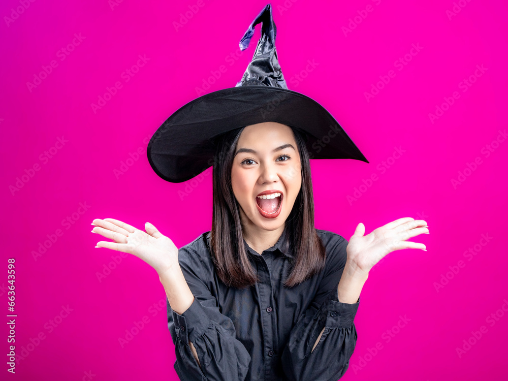 Portrait of an Asian Indonesian woman wearing a Halloween-themed costume with a witch hat, celebrating with clenched fists. Isolated against a magenta background. Portrait of an Asian Indonesian woman wearing a Halloween-themed costume with a witch hat, celebrating with clenched fists. Isolated against a magenta background.