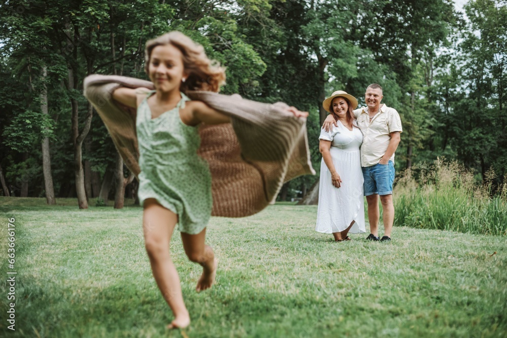 Obraz premium Lovely girl in fluttering knitted cardigan is running across lawn, smiling parents are watching. 