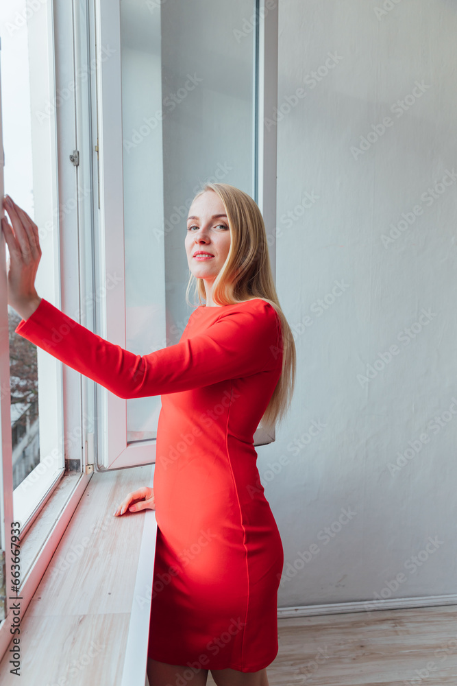 A woman in a red dress stands in a room by the window A woman in a red dress stands in a room by the window