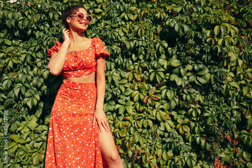 Young beautiful smiling hipster woman in trendy summer red dress. Sexy carefree woman posing in the street. Positive model outdoors. Cheerful and happy. Near green bushes. In sunglasses
