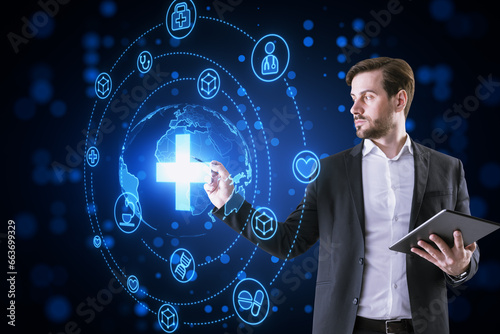 Attractive thoughtful young businessman with tablet using creative glowing blue medical hologram with cross and globe on dark background. Science, healthcare system and futuristic pharmacy concept.