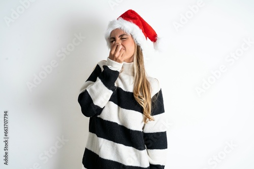 Beautiful hispanic woman wearing christmas hat and striped knitted sweater smelling something stinky and disgusting  intolerable smell  holding breath with fingers on nose. Bad smell