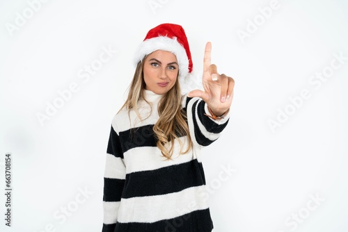 Beautiful hispanic woman wearing christmas hat and striped knitted sweater making fun of people with fingers on forehead doing loser gesture mocking and insulting.