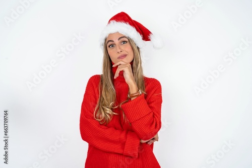 Beautiful hispanic woman wearing christmas hat and red knitted sweater looking confident at the camera smiling with crossed arms and hand raised on chin. Thinking positive.