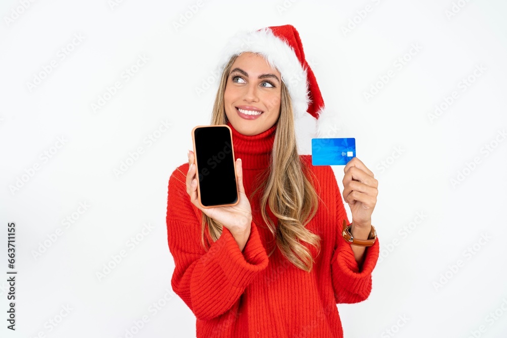 Beautiful hispanic woman wearing christmas hat and red knitted sweater holding bank card modern device looking empty space