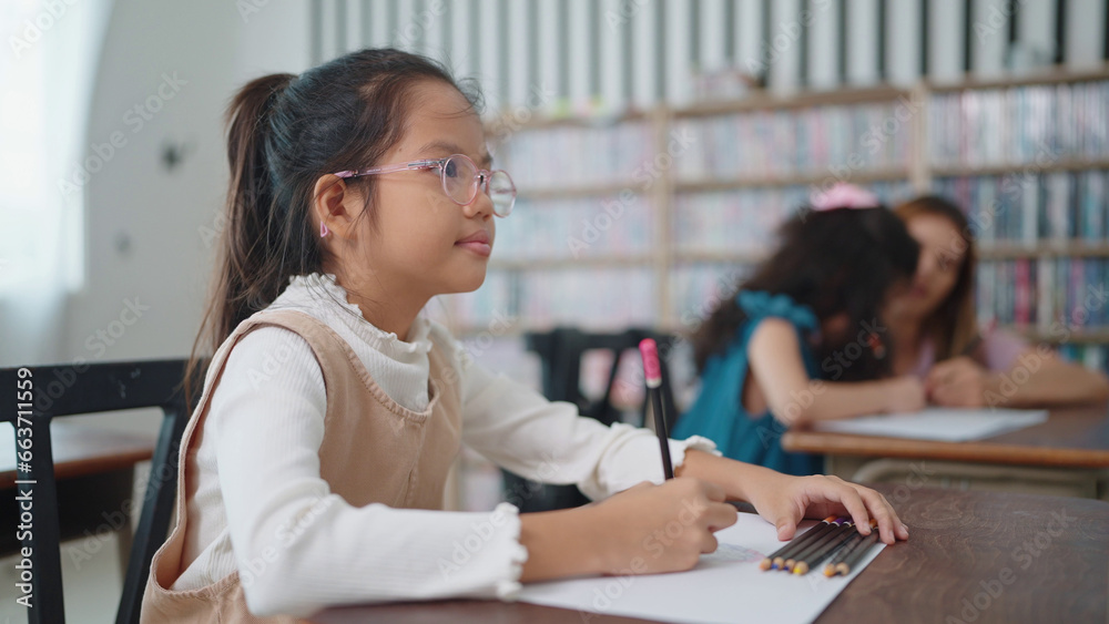 Obraz premium Asian school girl in glasses studying, writing notes in notebook at classroom. Schoolgirl studying and preparing for exams. little pupil writing at desk in classroom. Education knowledge concept