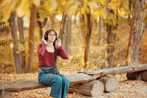 Red-haired teenage girl listens to music on headphones on a bench in an autumn park. Autumn season, yellow foliage.