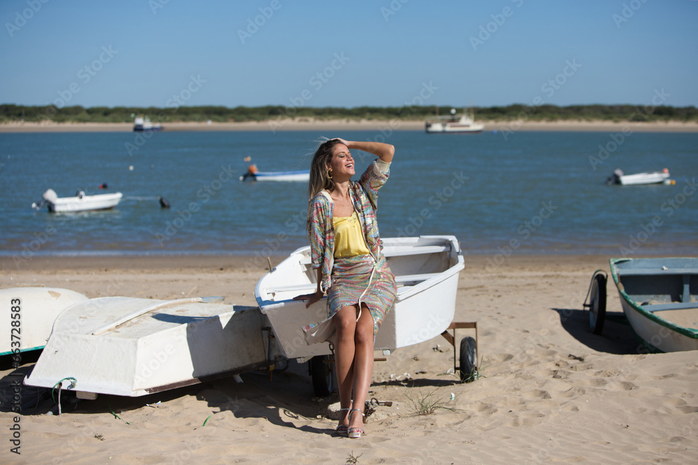 Obraz premium Pretty young blonde woman sitting in the fishermen's boats on the seashore. In the background on the horizon the blue sea and the boats working in the sea. Holiday and travel concept.