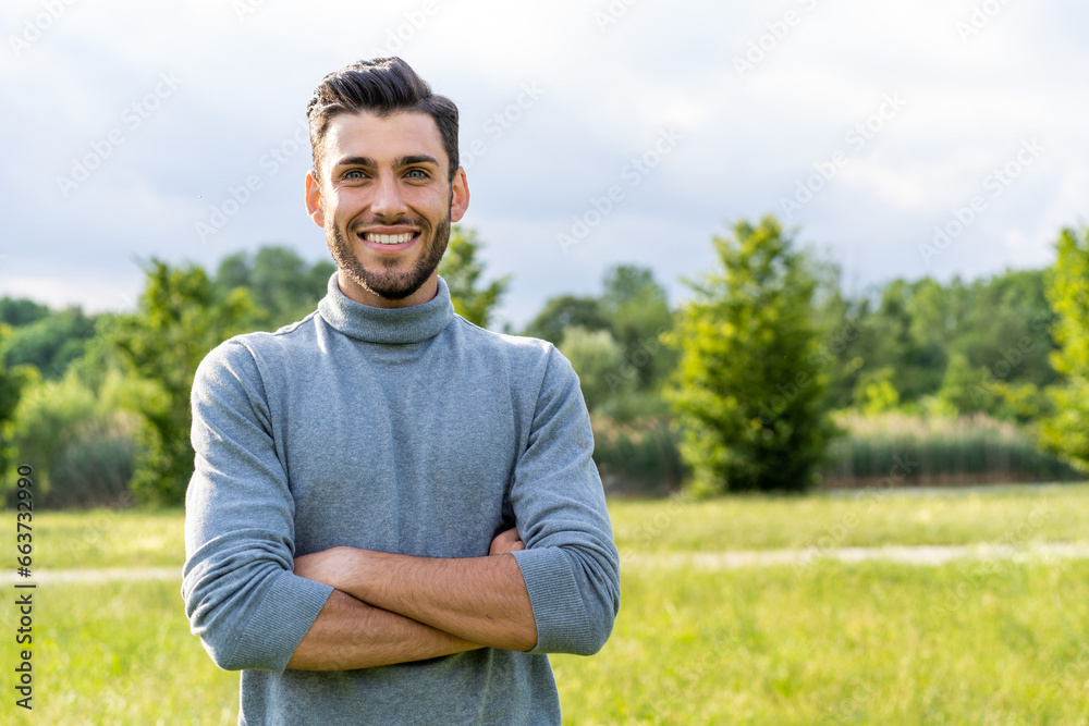 A handsome young Caucasian man with a beard stands confidently in an outdoor park, arms crossed. He offers a genuine smile, making direct eye contact with the camera.