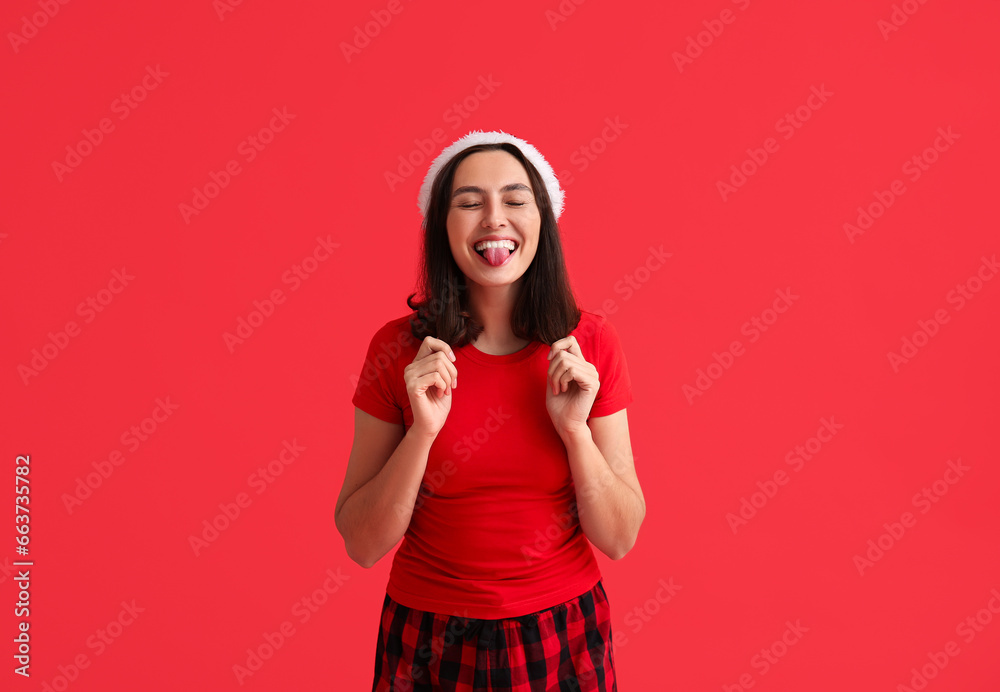 Beautiful young woman in pajamas and Santa hat on red background Beautiful young woman in pajamas and Santa hat on red background