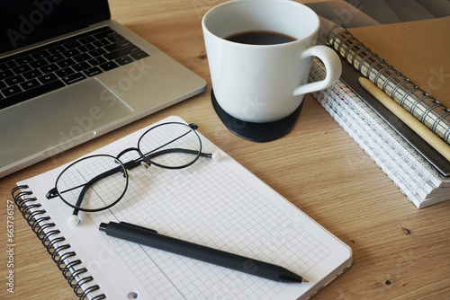 Notepad with pen and glasses, folders, open laptop and coffee cup nearby on wooden table