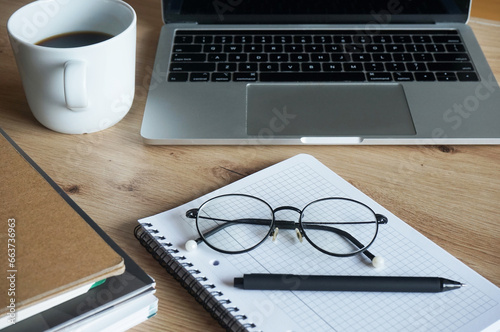 Notepad with pen and glasses, folders, open laptop and coffee cup nearby on wooden table