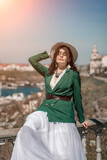 Woman walks around the city, lifestyle. A young beautiful woman in a green jacket, white skirt and hat is sitting on a white fence with balusters overlooking the sea bay and the city.
