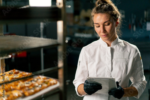 Portrait of a focused girl baker who checks the number of baked puffs in the bakery against list in a tablet 